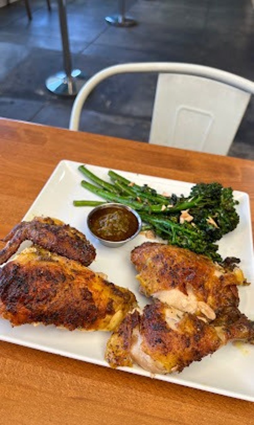 Grilled chicken pieces, broccolini, and a small dish of sauce on a white plate.